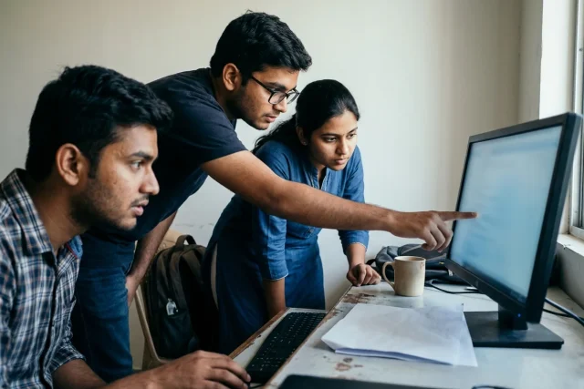 Three students grouped around one monitor, one of them pointing at the screen