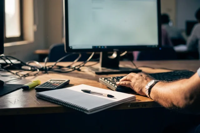 A desk close-up: an open ruled notebook and a pen beside a keyboard, with a calculator to one side
